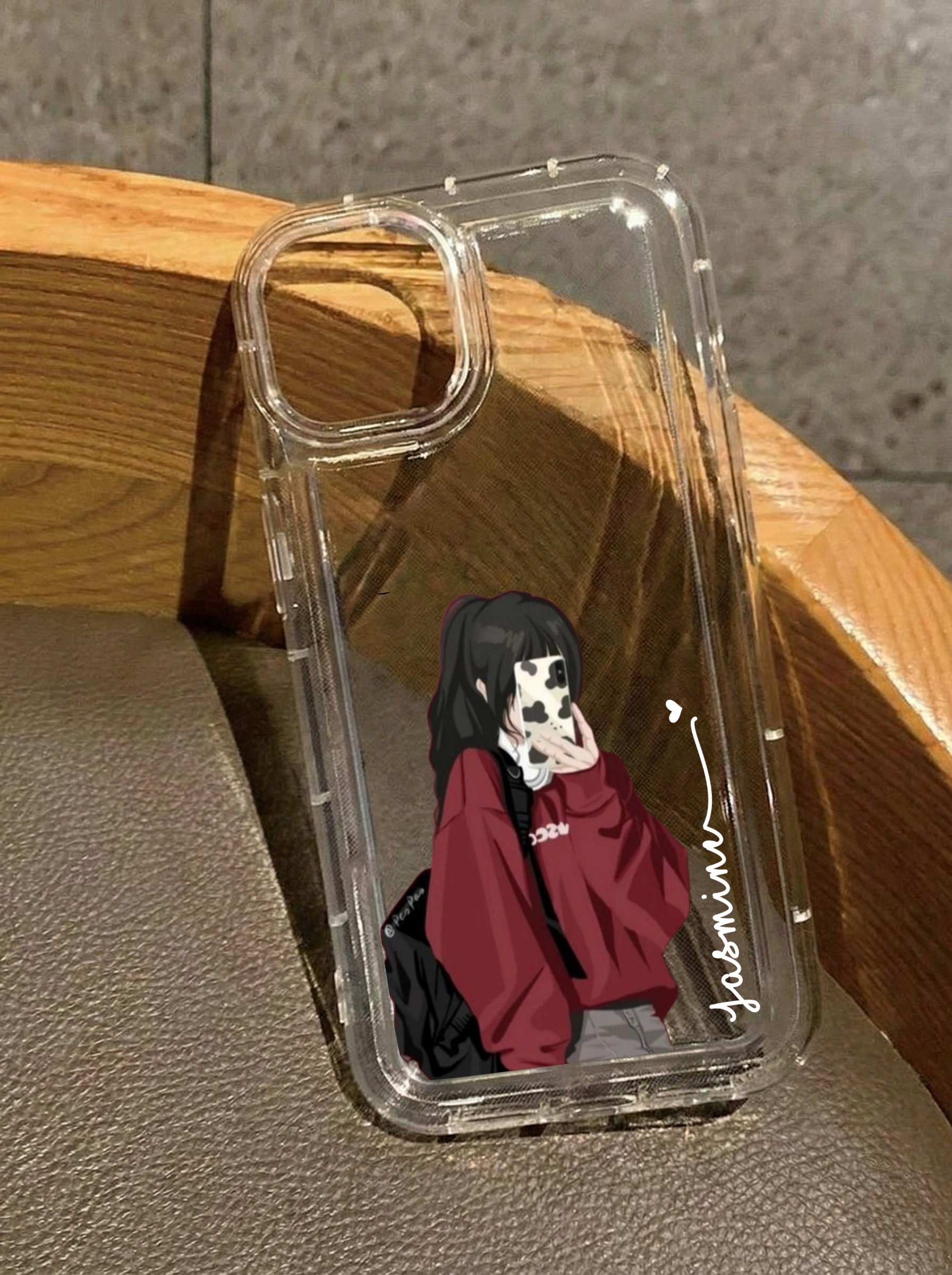Clear iPhone 16 case displaying an illustration of a girl in an oversized red hoodie and black backpack taking a selfie, customized with the name Jasmine in vertical white text.
